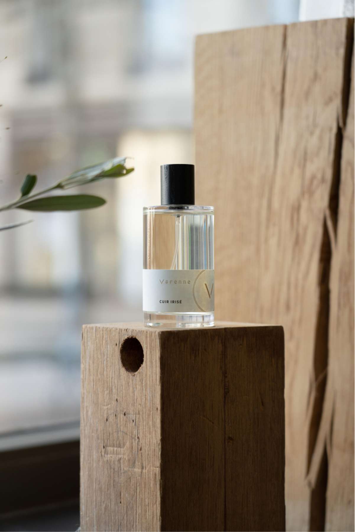 Photo parfum flacon verre avec étiquette sur laquelle figurent Varenne, son logo et le nom cuir irisé. Il est posé sur pied en bois dans vitrine avec un branchage